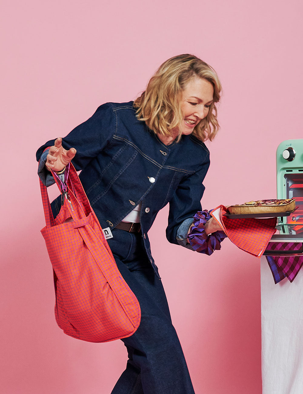 A woman in a dark denim jumpsuit holds a bright coral-red textured tote bag in one hand and a pizza in the other, posed against a solid pink studio background.