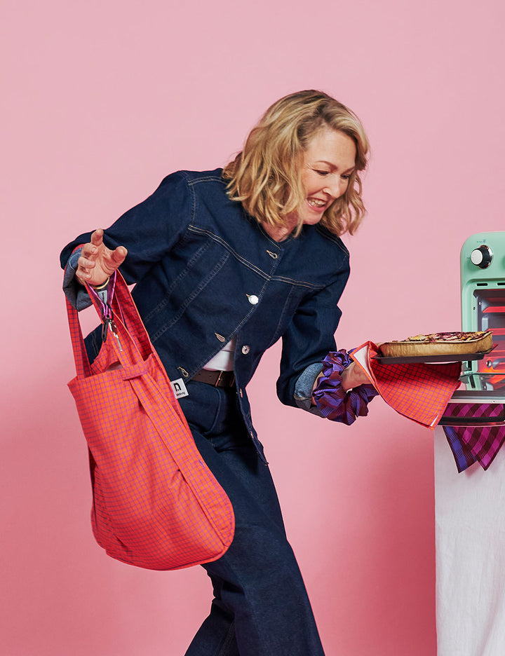 A woman in a dark denim jumpsuit holds a bright coral-red textured tote bag in one hand and a pizza in the other, posed against a solid pink studio background.