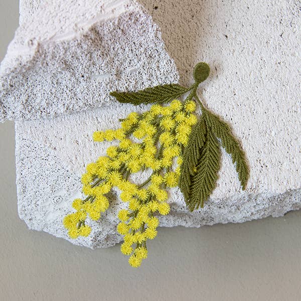 Botanopia mimosa embroidery brooch lying on a rock with a white backdrop.