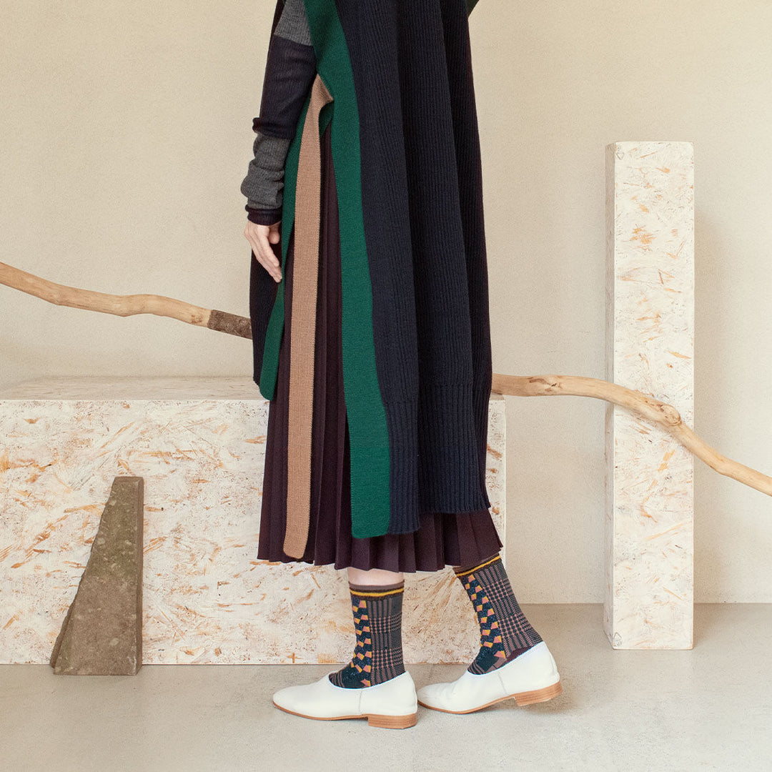 A model wearing Antique Patch socks in dark green, styled with a brown and navy outfit and white shoes, showcasing the geometric patterns of the socks.