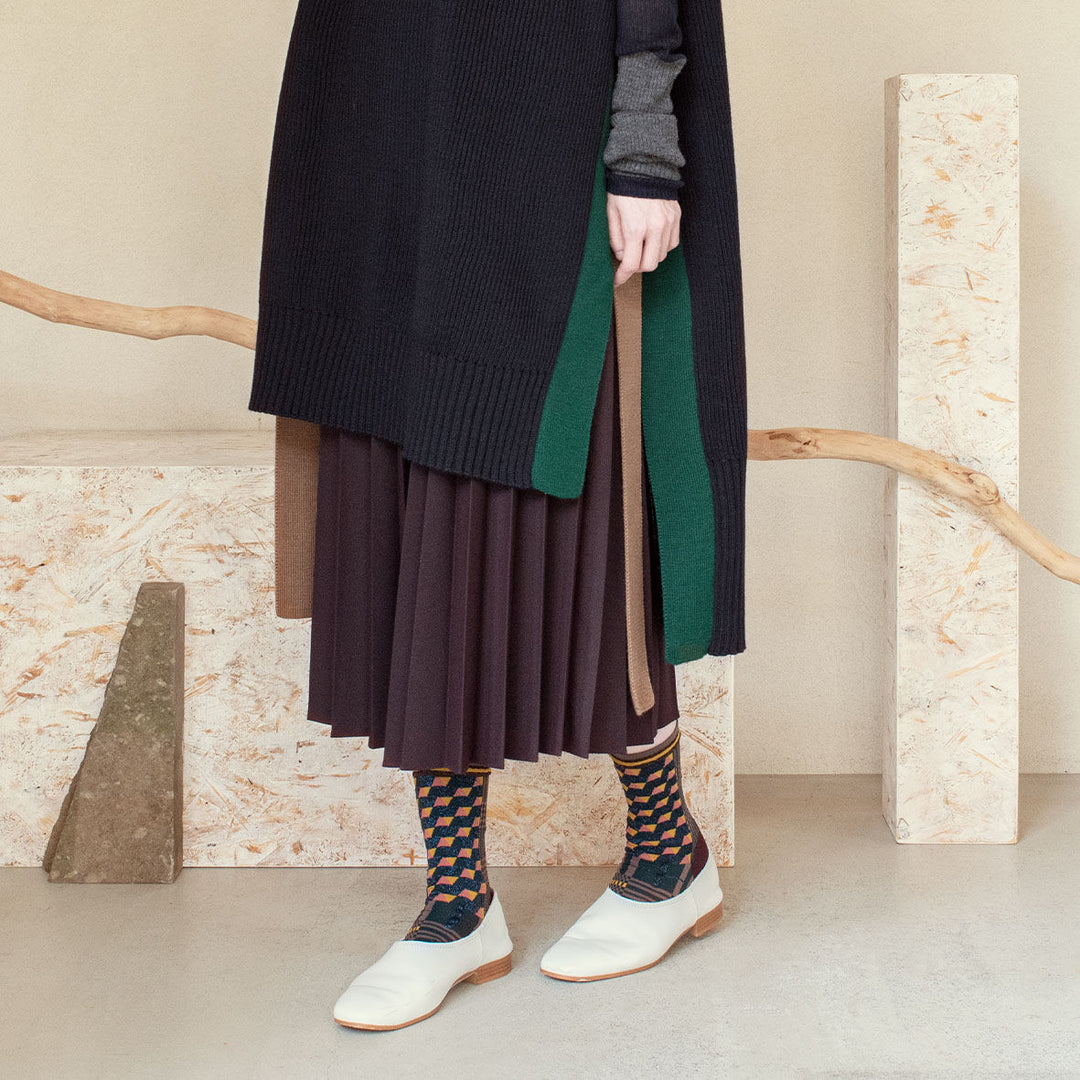 A model wearing Antique Patch socks in dark green, styled with a brown and navy outfit and white shoes, showcasing the geometric patterns of the socks.