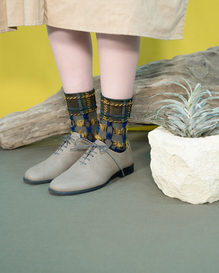 A model wearing Atelier Textiles Yellow socks in blue, yellow, and grey, made in Japan, paired with light leather shoes, showcasing their unique patterns and stylish look.