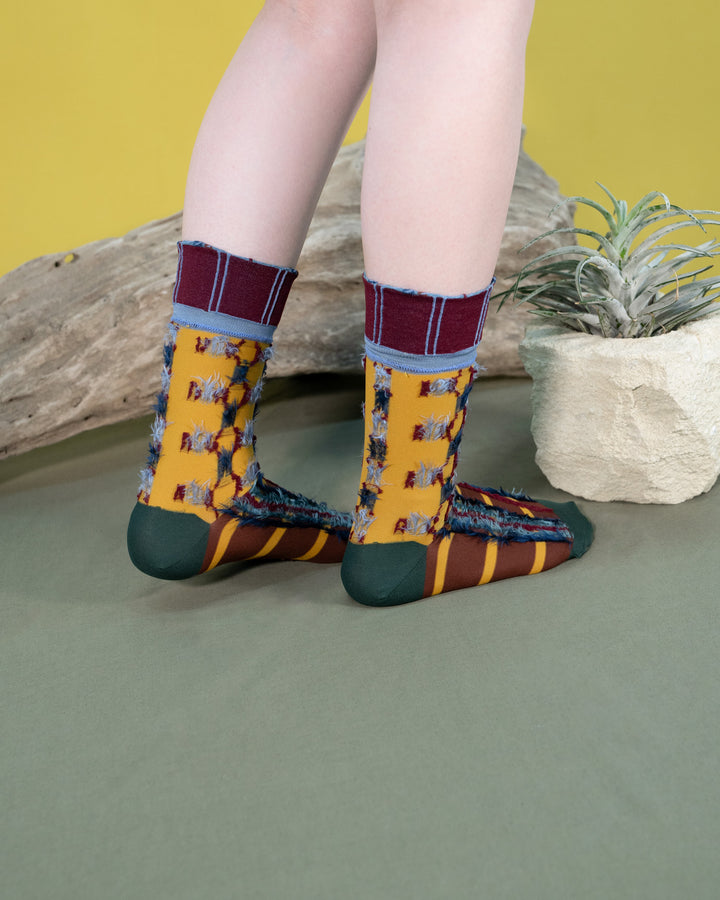 A model wearing Atelier Textiles Yellow socks, standing against a backdrop of vibrant plants, showcasing the socks' unique patterns and versatility.