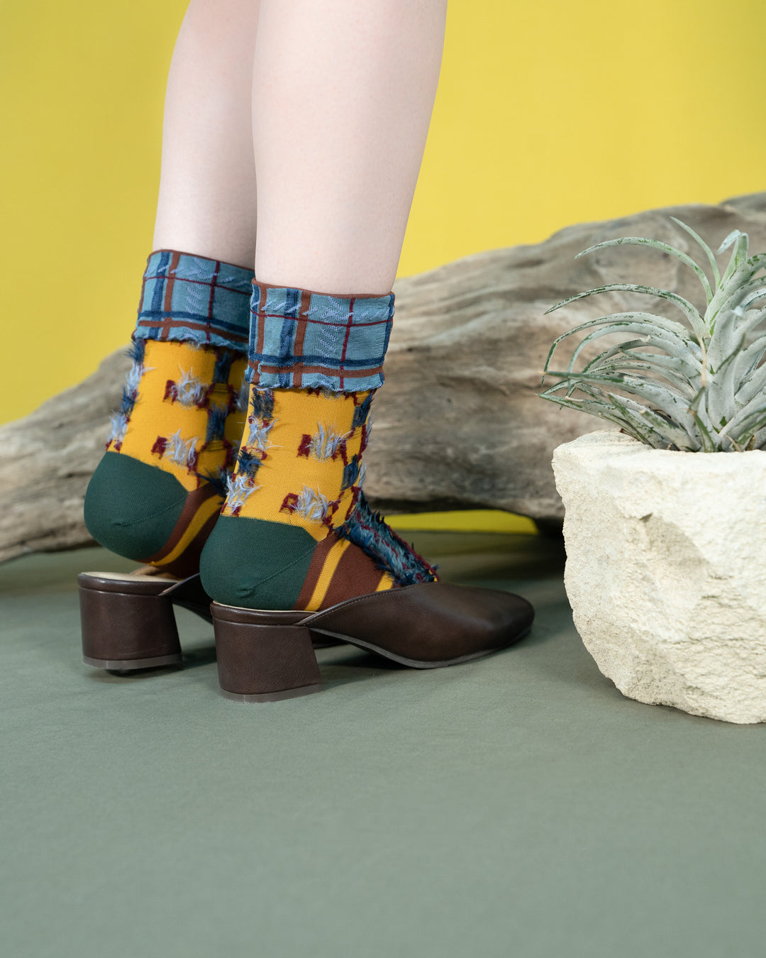 A model wearing Japanese Atelier Textiles Yellow socks, standing against a backdrop of vibrant plants, showcasing the socks' unique patterns and versatility.