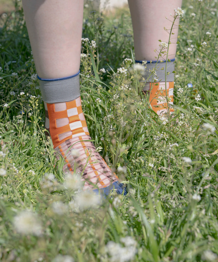 A model wearing COQ Picnic Window Socks in orange, made in Japan, standing on the grass, showcasing the checked pattern and translucent design.