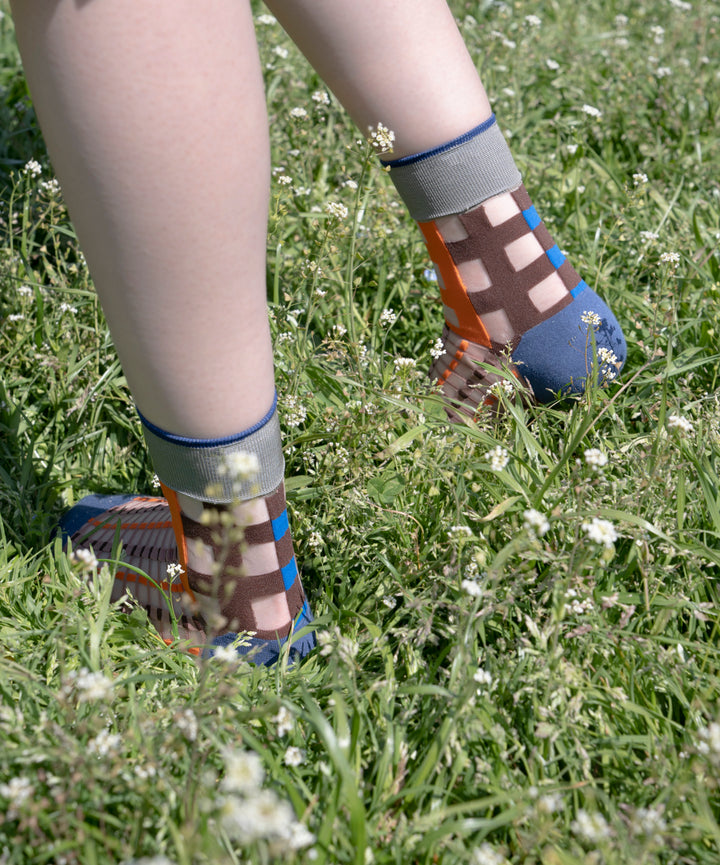 A model wearing COQ Picnic Window Socks in orange, made in Japan, standing on the grass, showcasing the checked pattern and translucent design.