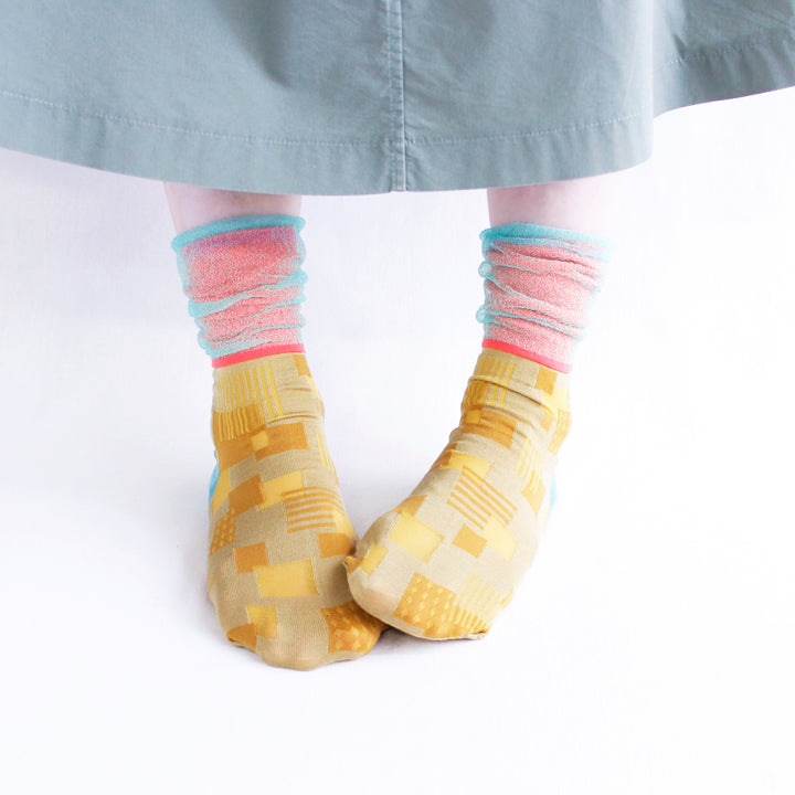 A model wearing COQ Sunlight Block Socks in yellow, made in Japan, styled with a grey dress, showcasing the unique geometric patterns of the socks.