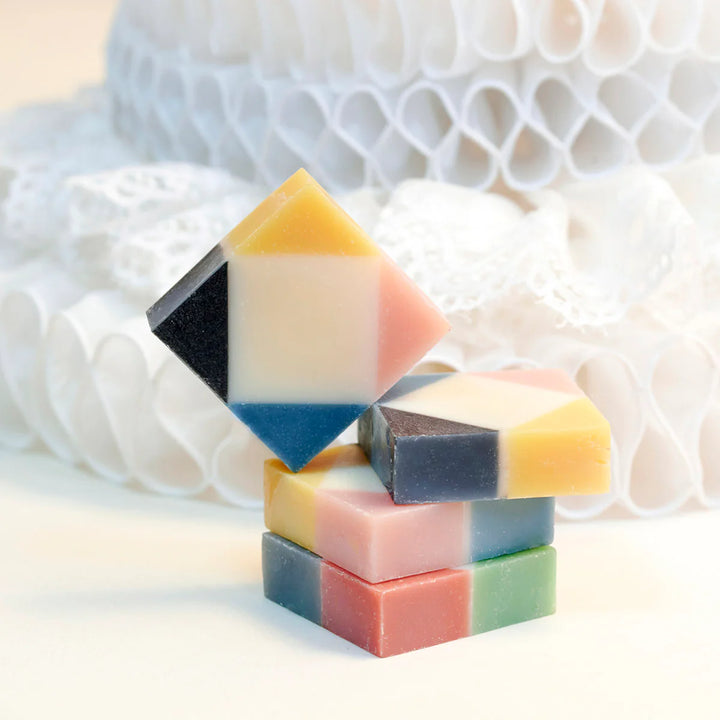 Four colorful unwrapped organic Ciment Arlequin soaps stacked, featuring rose and poudrée scents.