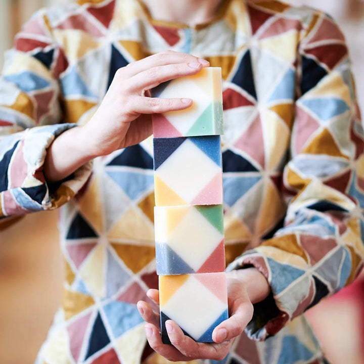 Person dressed as an Arlequin holding and stacking four colorful organic Ciment Arlequin soaps with rose and poudrée scents.