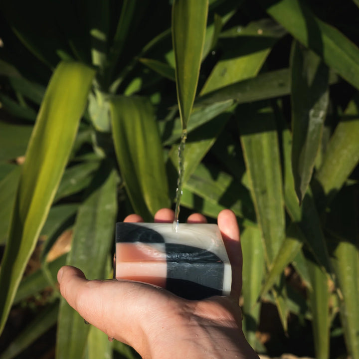 An organic Ciment Kimono Fleur de Cerisier soap held by a hand, with water dripping from leaves in the background.