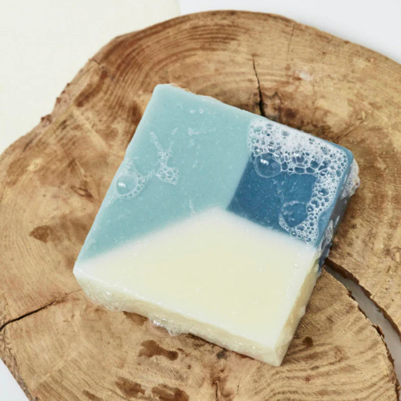 Unwrapped blue organic Ciment soap called Soap Window Fleurs de Pommier with an apple blossom scent, placed on a wet wooden platform with bubbles around it.