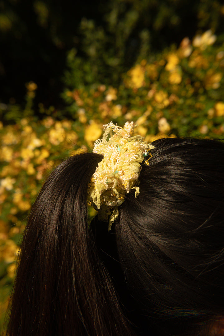 A person tying her ponytail with a Lowland Summer confetti scrunchie in Piña Colada, against a yellow floral background.