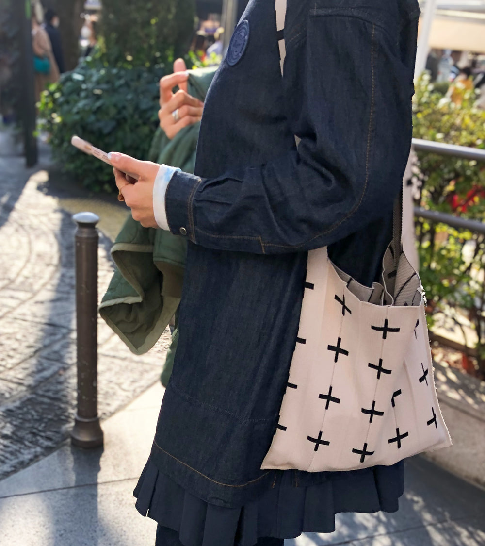 A model carrying the beige KNT 365 Knitty Tote Bag with a cross pattern over her shoulder on the street, paired with a denim jacket for a stylish look.