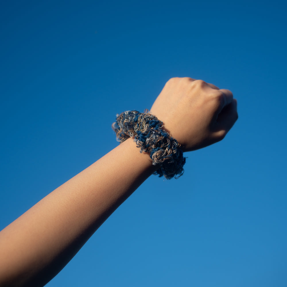 A hand wearing a handmade blue crochet scrunchie made from vibrant Japanese yarns with playful ruffles.