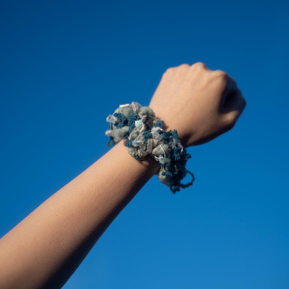 A hand wearing a handmade blue crochet scrunchie made from vibrant Japanese yarns with playful ruffles.