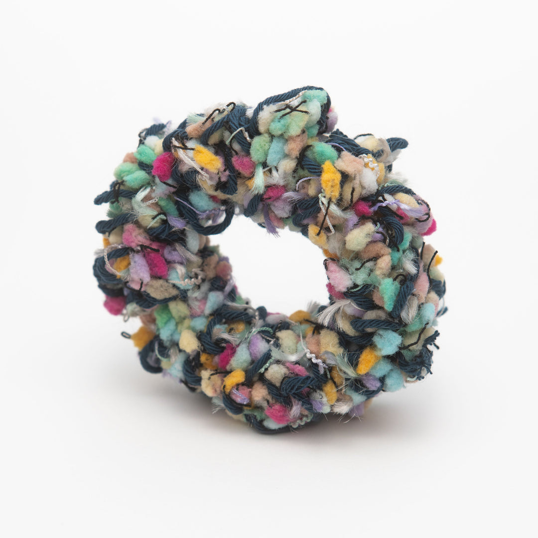 A handmade crochet scrunchie featuring a vibrant mix of colorful yarns with playful ruffles, styled as a wrist accessory.