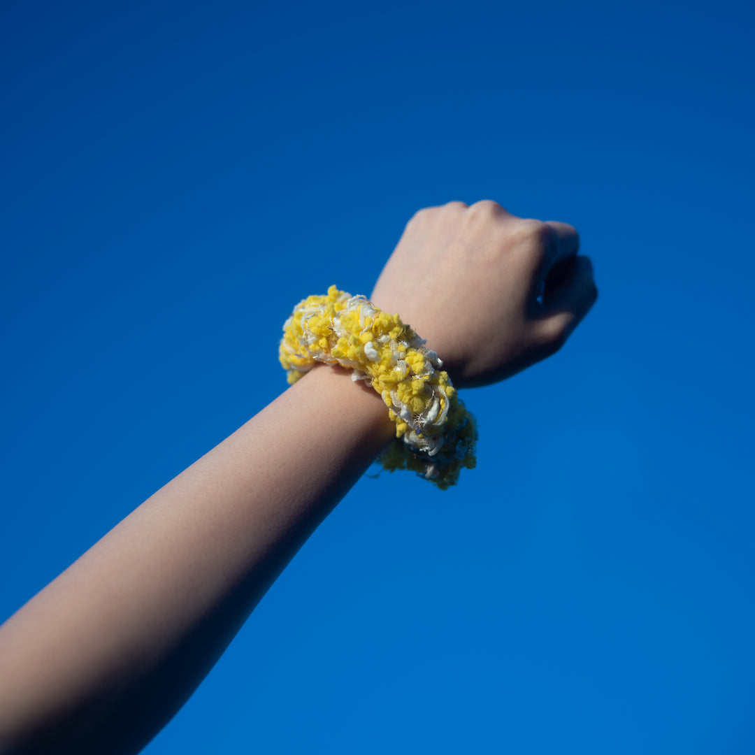 A hand wearing a handmade yellow crochet scrunchie made from vibrant Japanese yarns with playful ruffles.