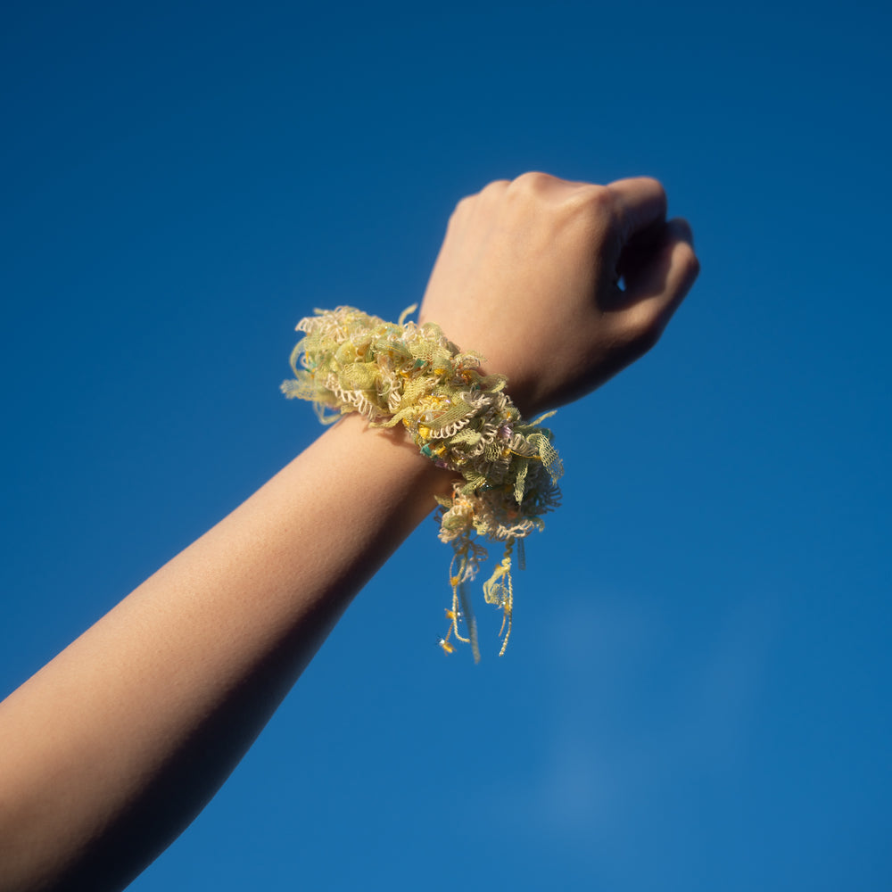 A hand wearing a handmade yellow crochet scrunchie made from vibrant Japanese yarns with playful ruffles.