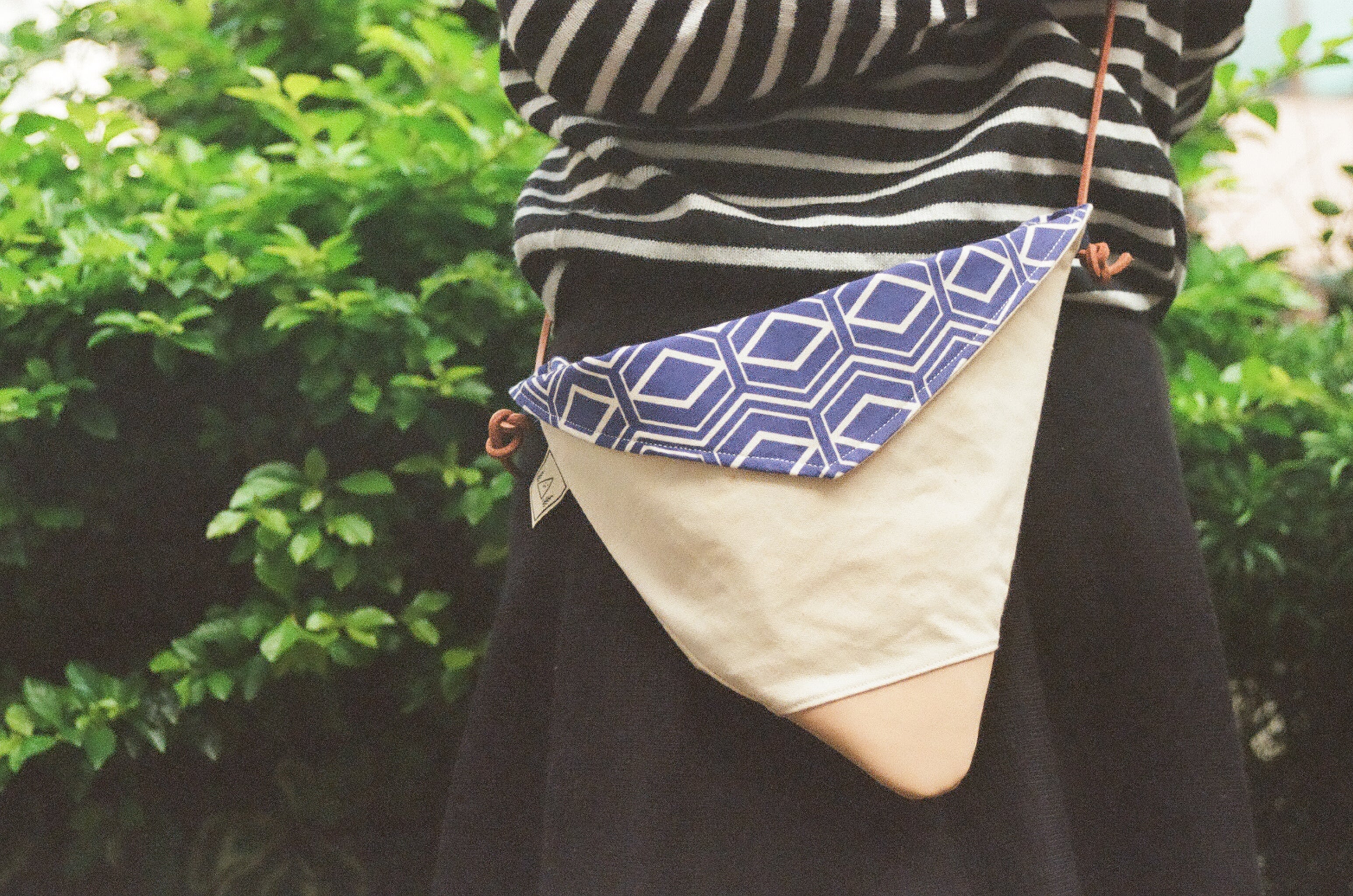 A product shot of a handmade triangular leather and fabric bag featuring a blue geometric pattern. The person wearing the bag stands against a green, natural backdrop.