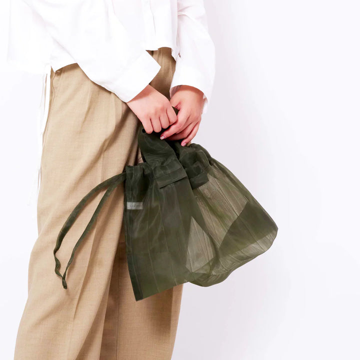 A model holding a khaki Japan Mate Mono see-through bag with her hands, styled to match her white shirt.