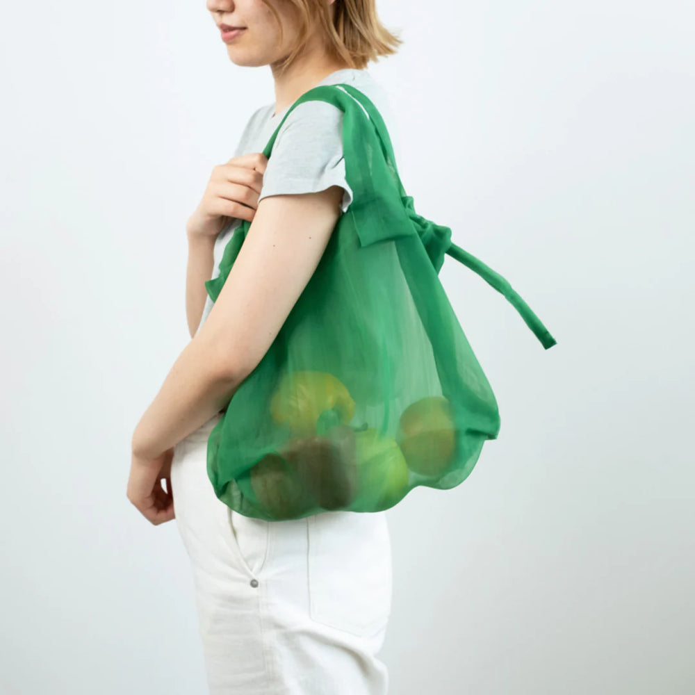 A model carrying a green Japan Mate Mono see-through bag on her shoulder, filled with fruits, showcasing its practicality for grocery shopping.