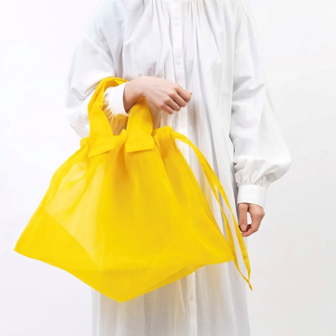 A model holding a yellow Japan Mate Mono see-through bag with her hand, styled to match her white minimal shirt.