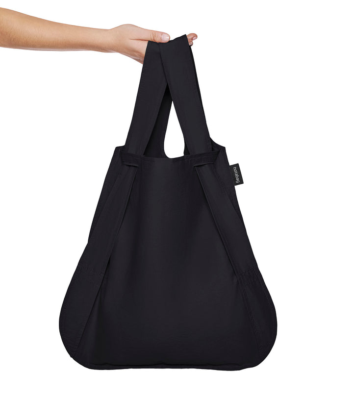 A model holding a black Notabag by its handle with her arm, showcasing its spacious capacity.