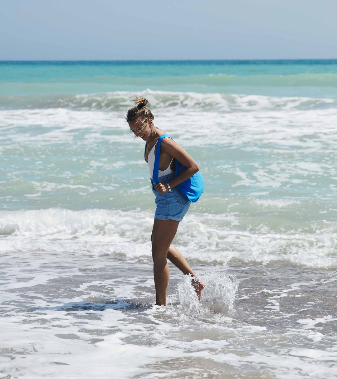 A model wearing a blue Notabag as a backpack, walking in the water on a beach, showcasing its stylish and functional design.