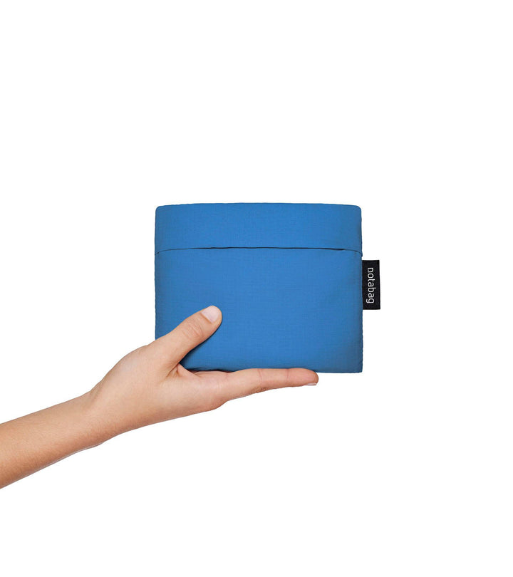 A model holding a folded blue Notabag in her hands, showing its compact size, similar to a wallet. The bag's slim, minimalist design is visible as she begins to unfold it, highlighting its portability and convenience for easy storage.