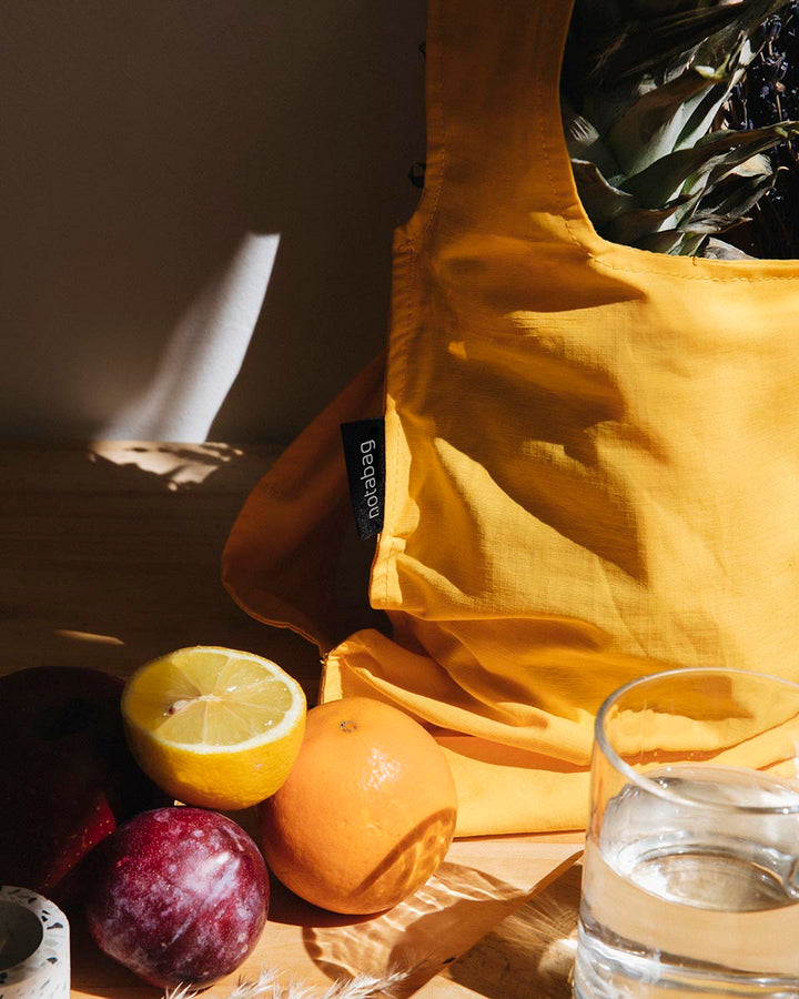 Image of the golden yellow Notabag displayed as a tote bag on a desk, accompanied by some fruits next to it, showcasing its suitability for grocery shopping.