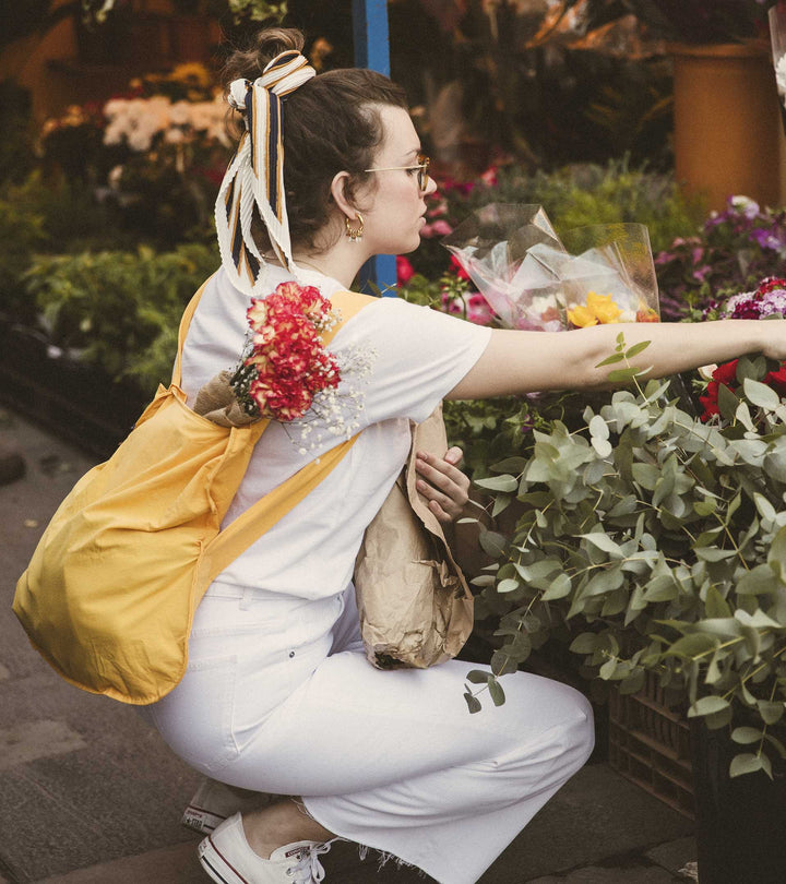 A model wearing the golden yellow Notabag as a backpack while heading to a flower market, highlighting its vibrant color and practicality.