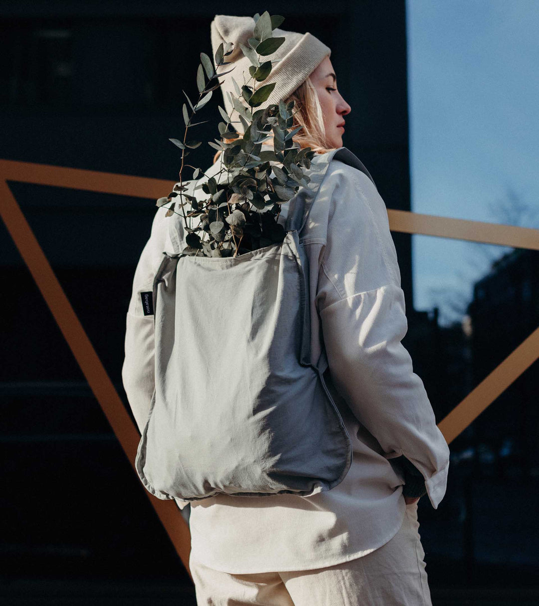 A model wearing a grey Notabag as a backpack, placing some plants inside while dressed in her winter outfit, highlighting the bag's stylish and functional design.