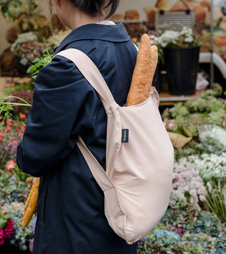 A model wearing a sand beige Notabag as a backpack while heading to a flower market, with a loaf of bread inside her bag, styled to match her navy winter jacket.