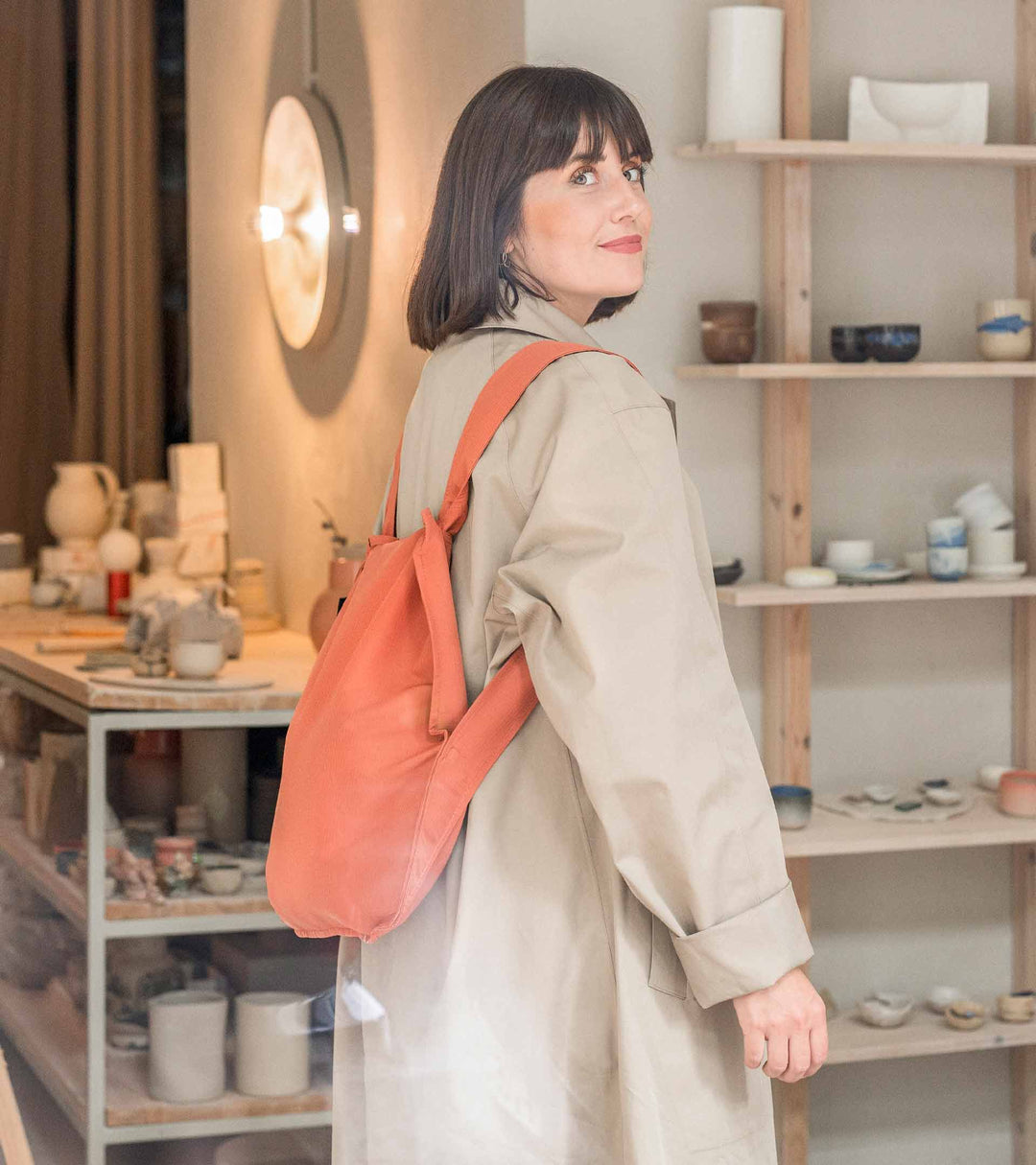 A model wearing a folded Terracotta Notabag as a backpack, paired with a long beige trench coat, standing in her ceramic studio.