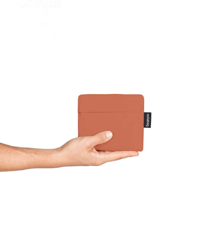 A model holding a folded Terracotta Notabag in her hands, showing its compact size, similar to a wallet. The bag's slim, minimalist design is visible as she begins to unfold it, highlighting its portability and convenience for easy storage.