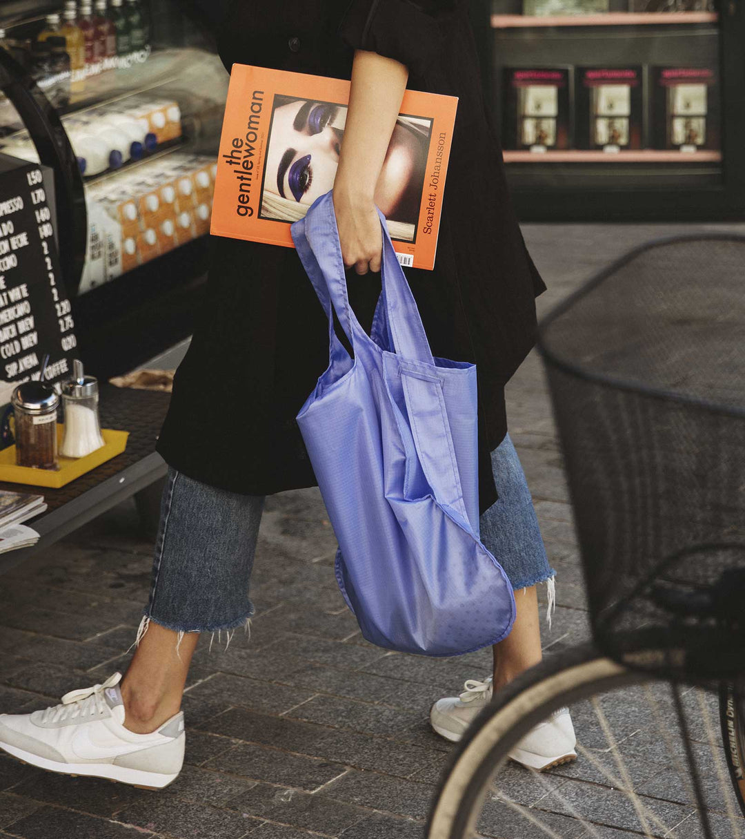 A model carrying a cornflower purple Notabag while shopping in a hipster area filled with cafes, showcasing the bag's stylish design and functionality.