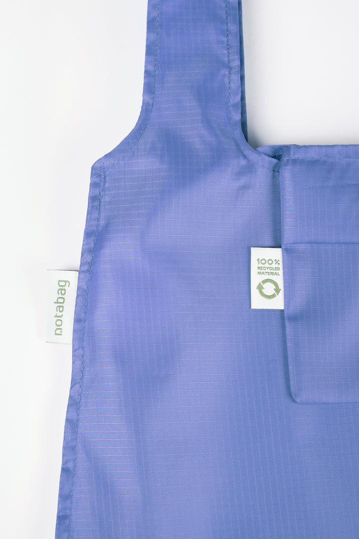 Close-up product shot of a cornflower purple Notabag, highlighting its texture and the label that states it is made from 100% recycled materials.