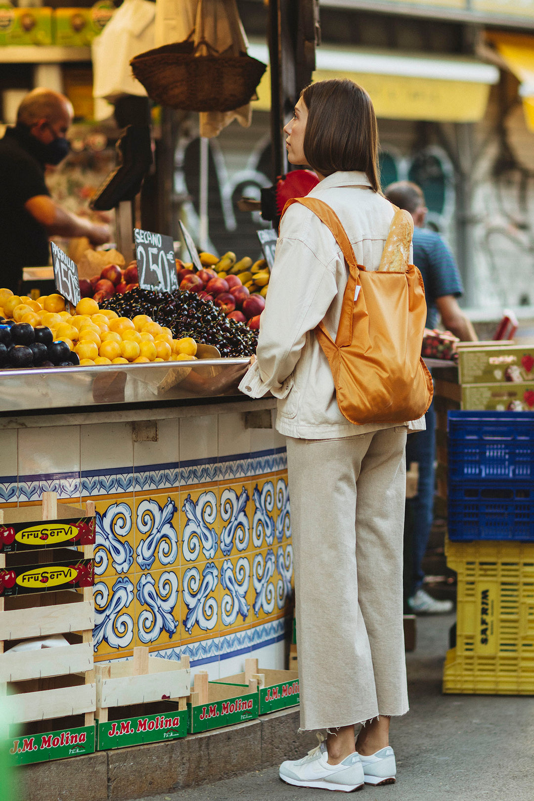 Woman wearing a Notabag as a backpack, walking through an outdoor fruit market filled with colorful produce and stalls.