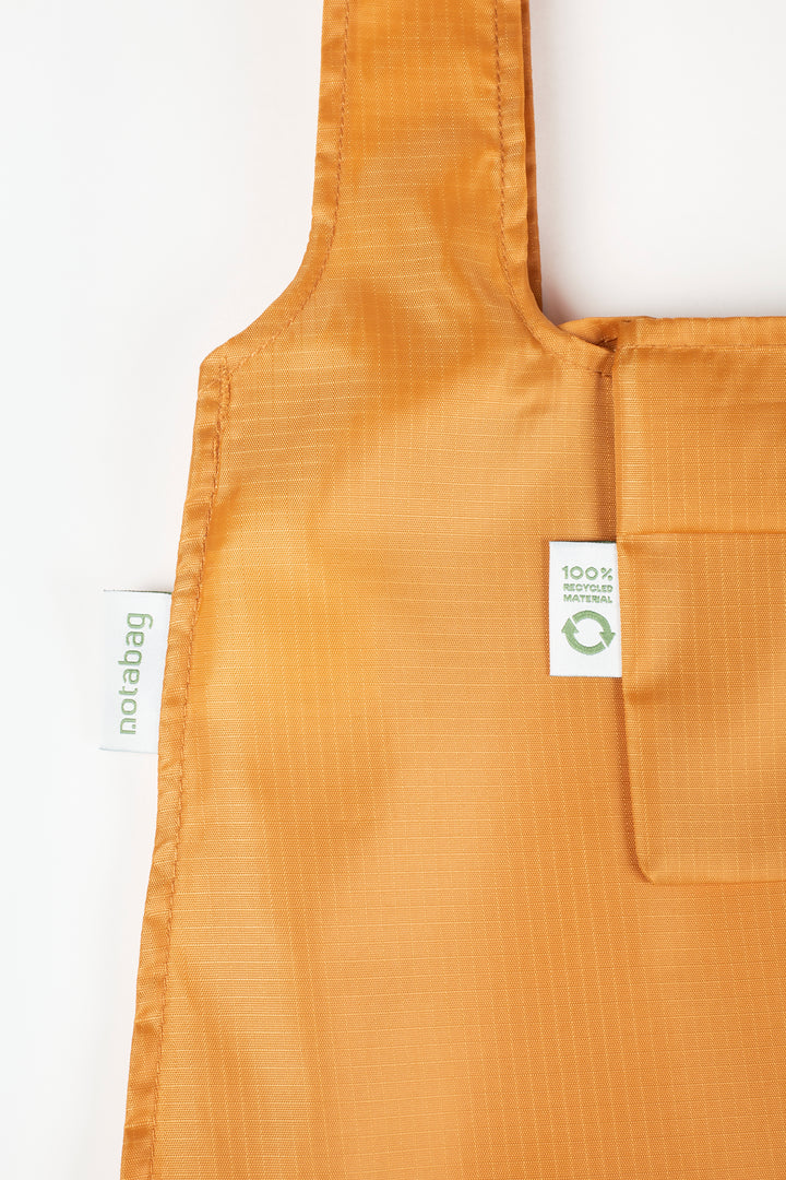 Close-up product shot of a mustard orange Notabag, highlighting its texture and the label that states it is made from 100% recycled materials.