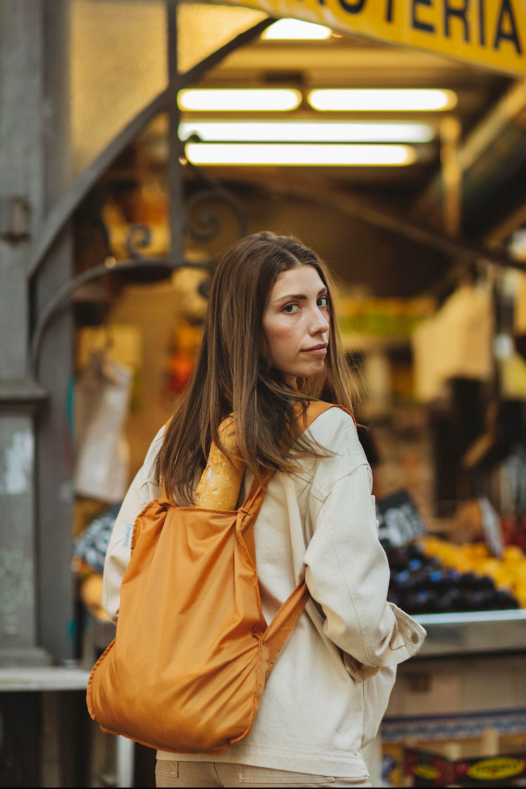 Woman wearing a Notabag with bread inside, styled to match her beige jacket as she strolls through a market or street.