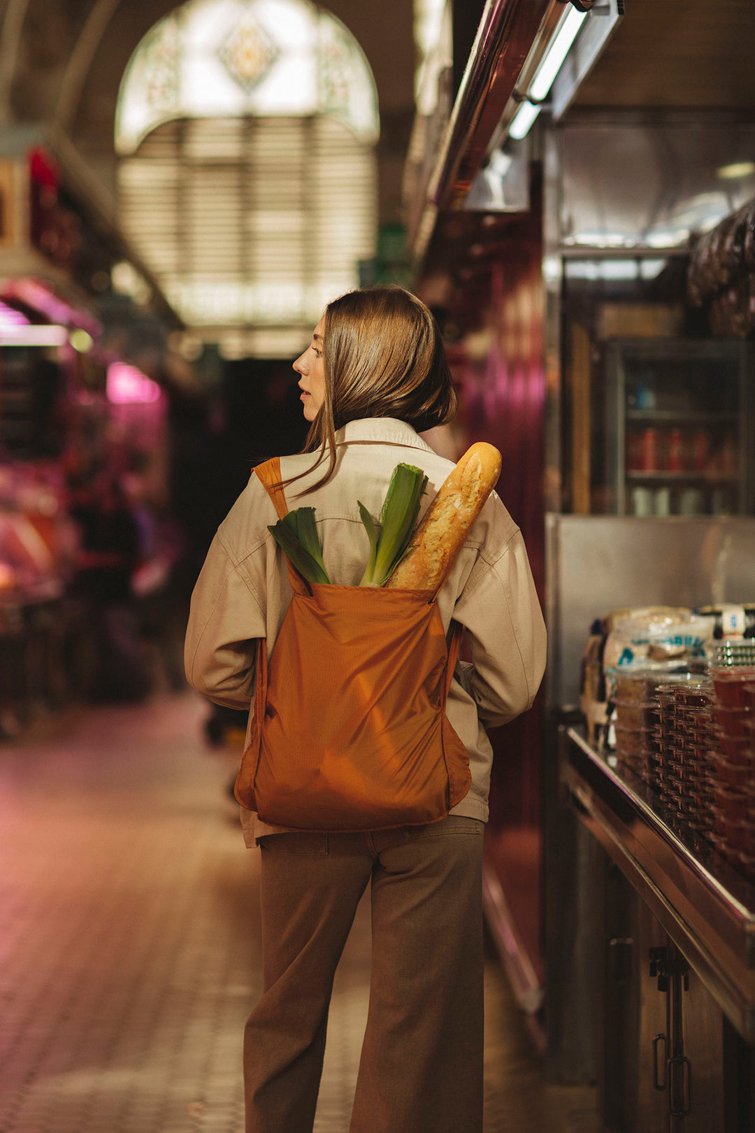 Woman wearing a mustard-colored Notabag as a backpack, filled with groceries while she shops at an outdoor market.