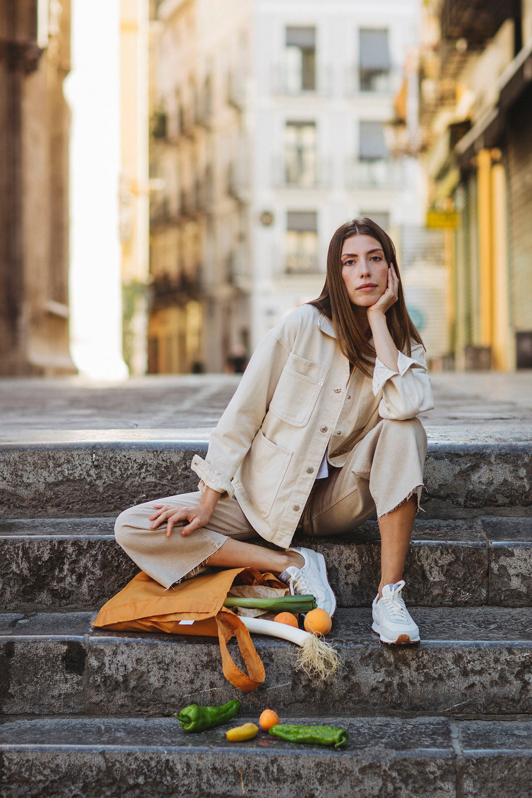 A woman sitting on the street facing the camera, with a Notabag beside her legs filled with groceries, looking relaxed and enjoying the moment.