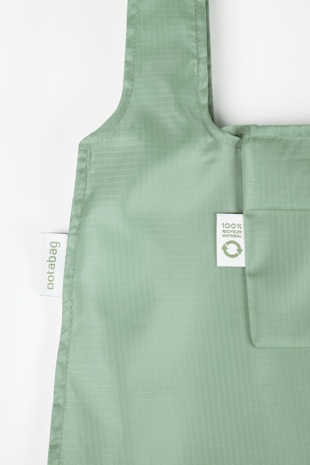 Close-up product shot of a sage green Notabag, highlighting its texture and the label that states it is made from 100% recycled materials.