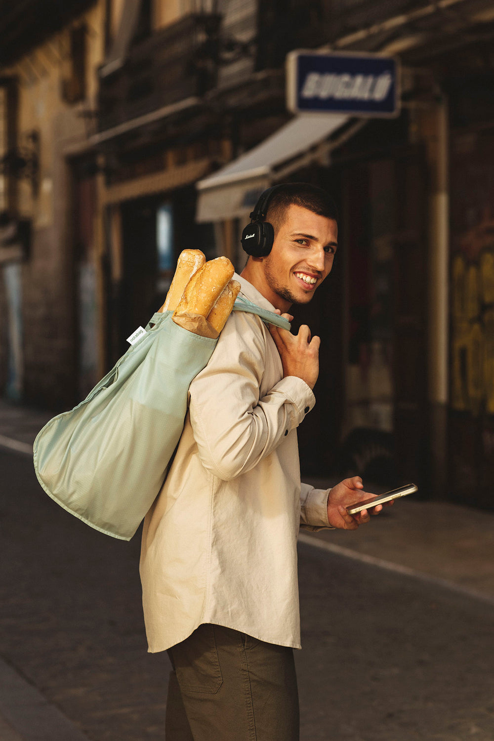 Man walking down the street with a sage green Notabag over his shoulder, a baguette sticking out, and holding a phone in his other hand.