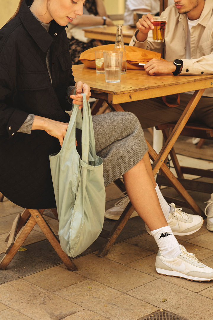 "A model seated at an outdoor café, reaching into her sage green Notabag to retrieve an item. The spacious, water-resistant bag sits open, showing it can hold numerous items, with the durable, textured fabric visible. Sunlight filters onto the bag, emphasizing its stylish yet practical design in the café setting.