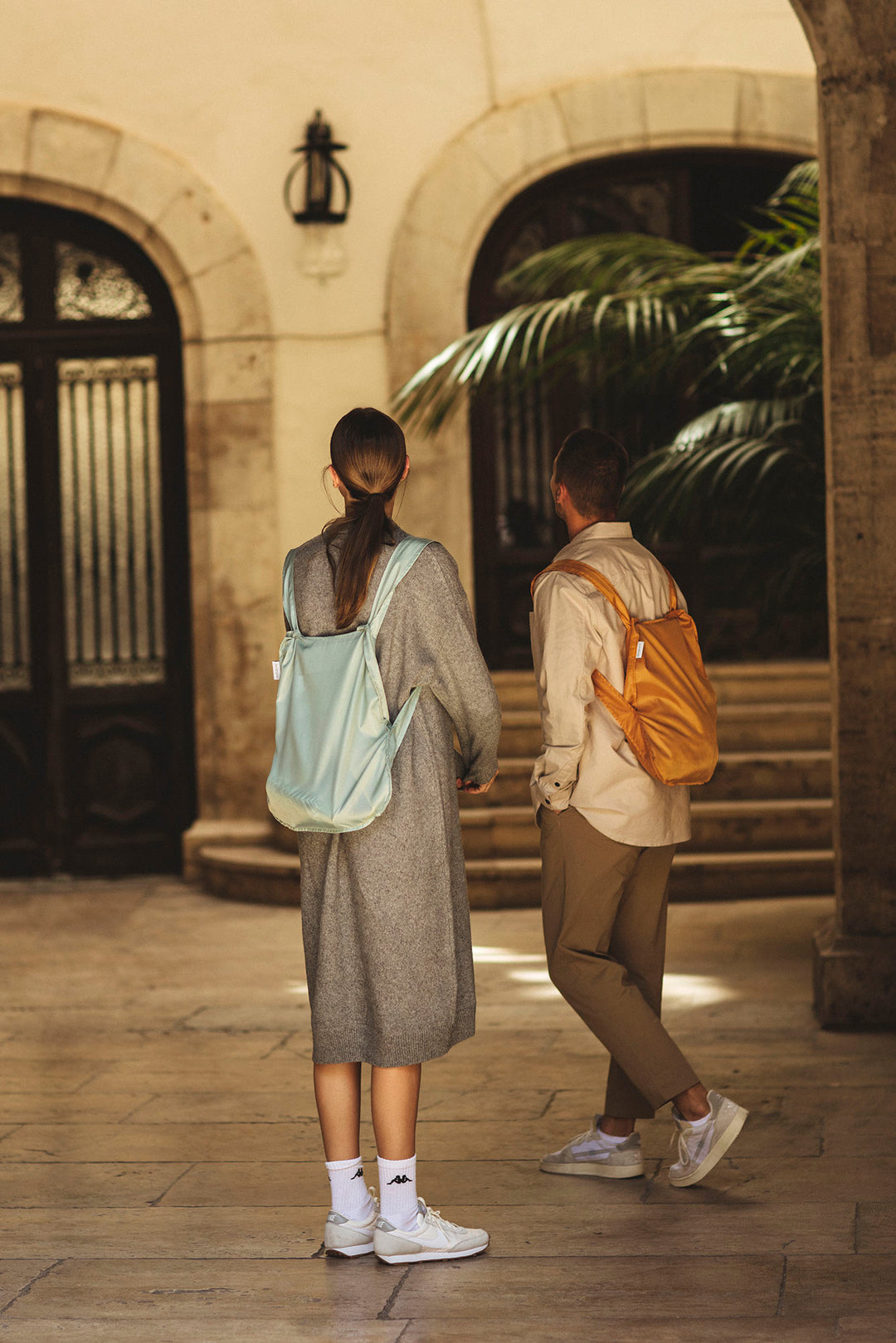 Two people wearing the recycled, water-resistant Notabag backpacks, walking together in a scenic, sunlit plaza with Spanish-style architecture in the background, featuring terracotta roofs, archways, and cobblestone paths.