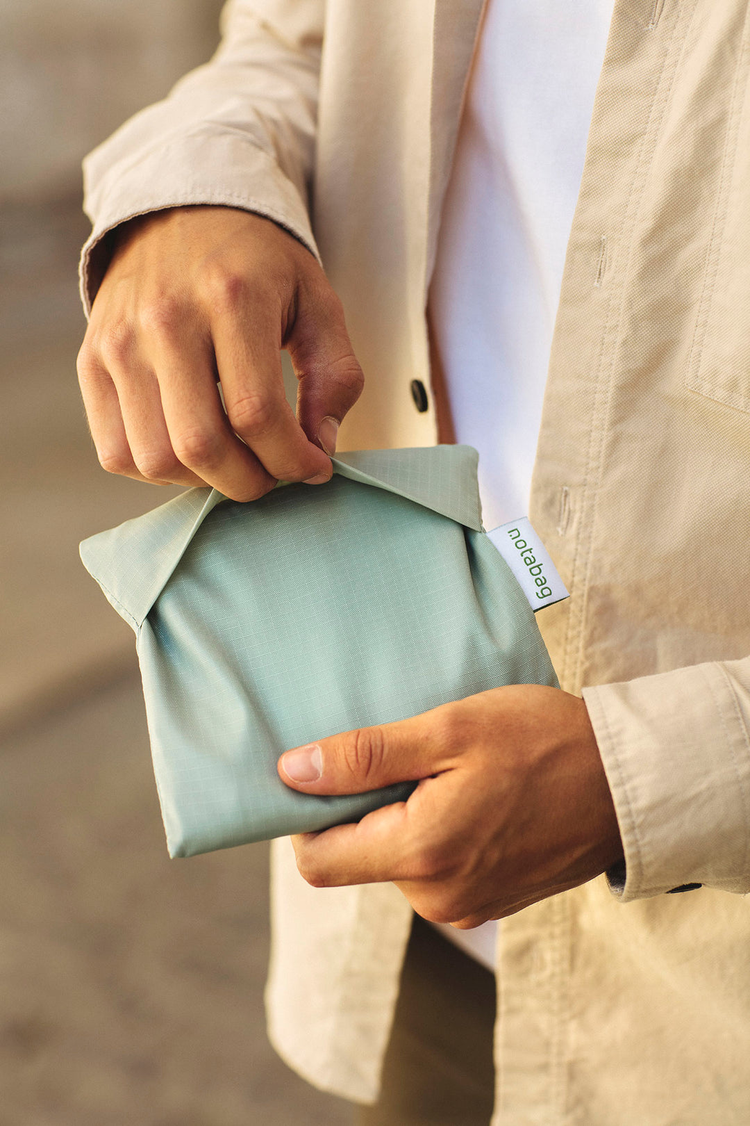 A model holding a folded sage green Notabag in her hands, showing its compact size, similar to a wallet. The bag's slim, minimalist design is visible as she begins to unfold it, highlighting its portability and convenience for easy storage.