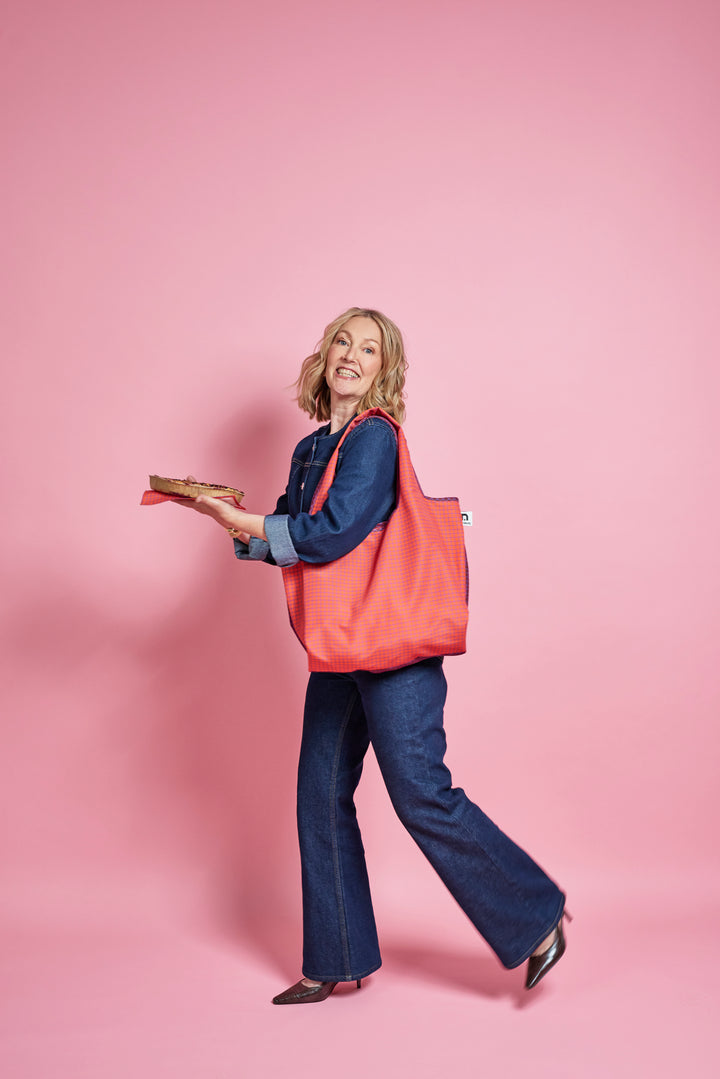 A woman in a blue denim outfit carrying a large coral-pink tote bag over her shoulder, holding a book in her other hand, set against a solid pink studio background.