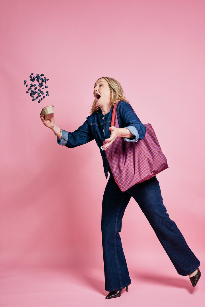 A woman in a denim outfit and heels is captured mid-motion against a solid pink studio background, holding a small cup with blueberries floating above her head. She is carrying a large magenta-and-white checkered tote bag.
