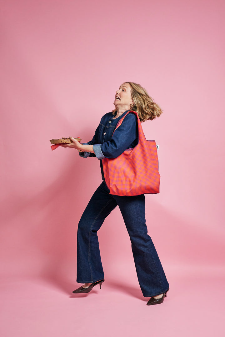 A woman in a blue denim outfit carrying a large coral-red tote bag over her shoulder, posed against a solid pink studio background.